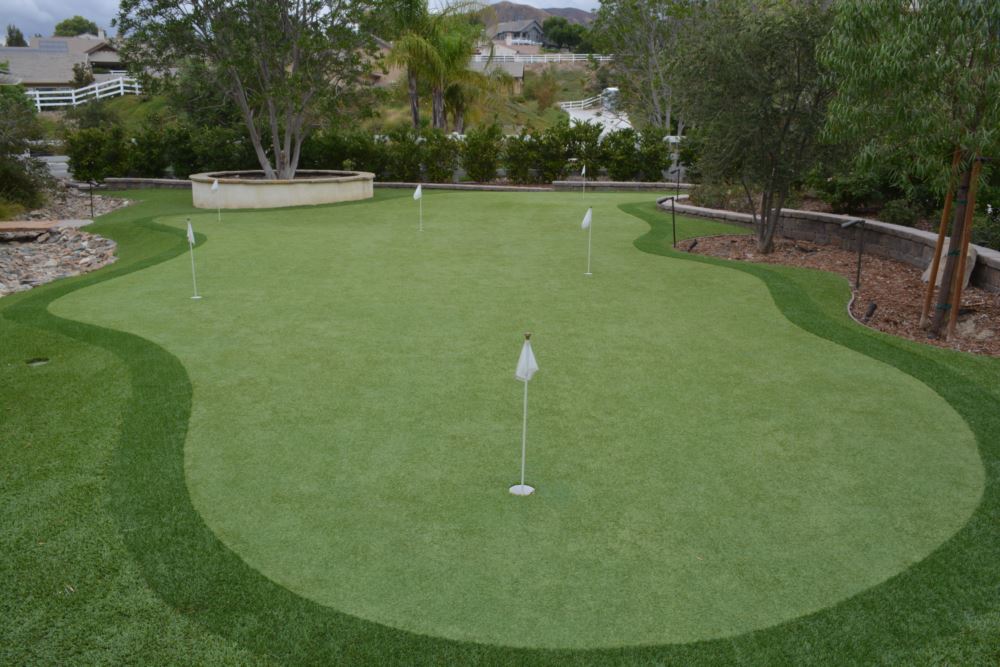 Custom backyard synthetic turf putting green with multiple flagsticks, contoured design, and realistic fringe, surrounded by landscaped trees and retaining walls for year-round golf practice.