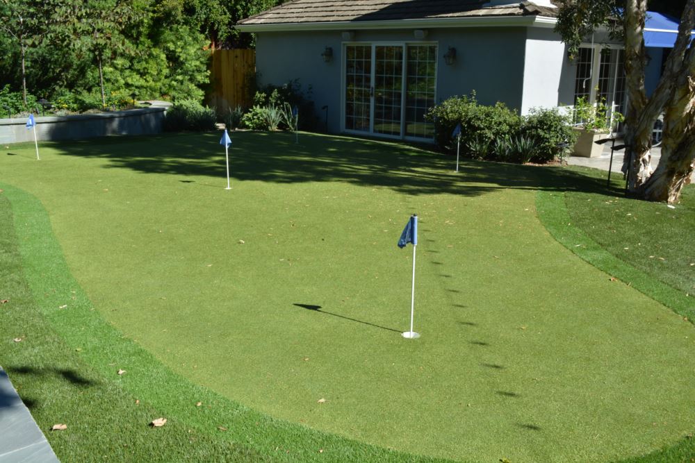 Residential backyard synthetic turf putting green with multiple practice holes and realistic fringe, installed near a patio and garden landscaping for year-round golf practice at home.