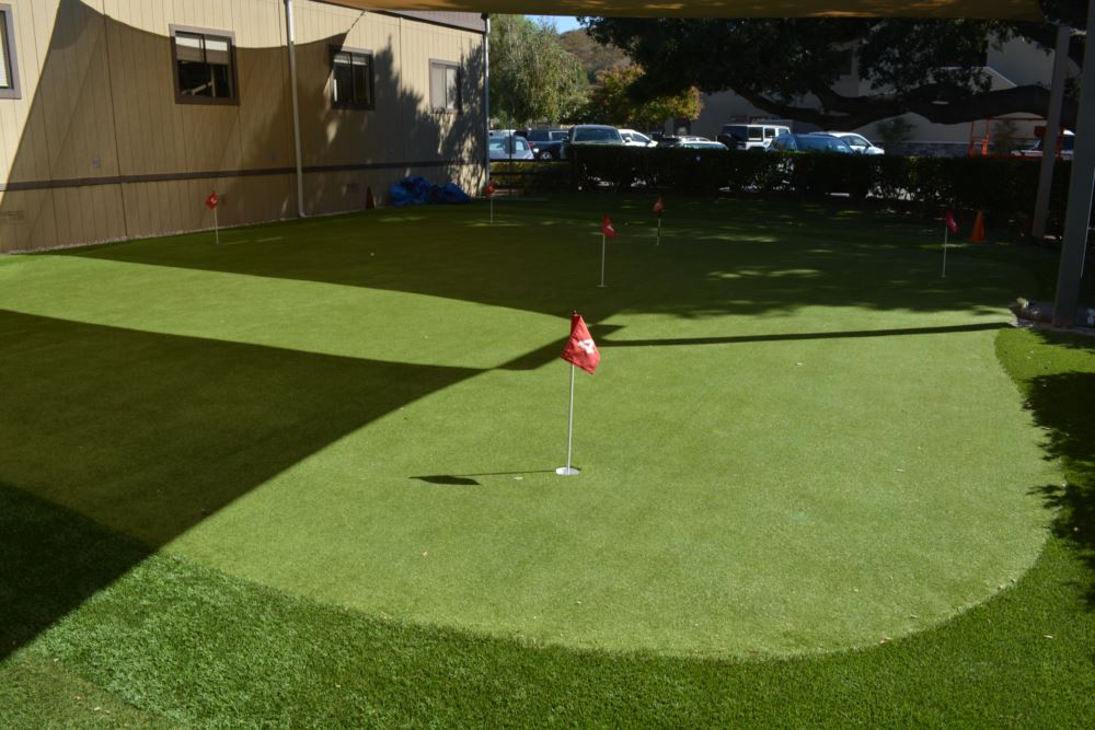 Artificial putting green with multiple numbered red flag pins and distinct turf speed zones installed beneath a shade sail at a commercial facility