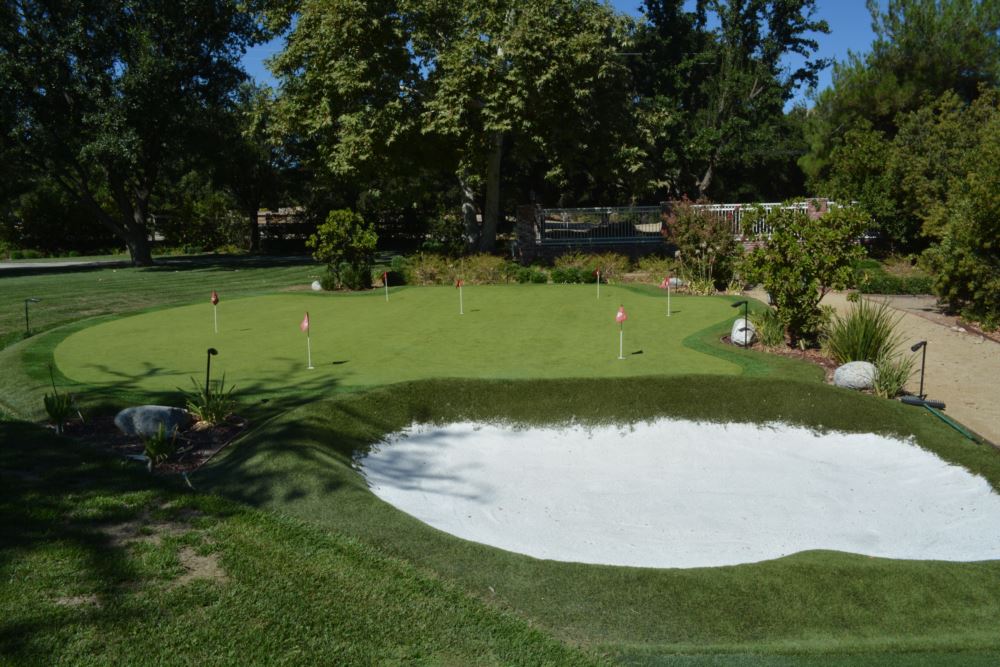 Backyard golf putting green with white sand bunker, multiple practice holes, and red flags surrounded by lush trees and ornamental plantings